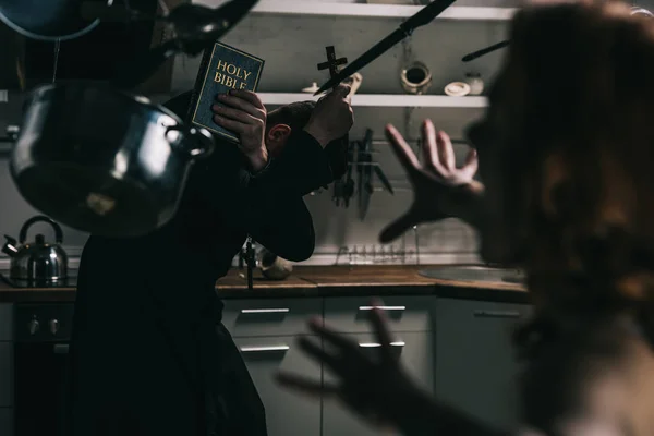 Selective focus of evil demon with levitating cookware and exorcist with cross and bible in kitchen — Stock Photo