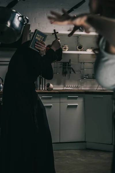 Selektiver Fokus des bösen Dämons mit schwebendem Kochgeschirr und Exorzist mit Kreuz und Bibel in der Küche — Stockfoto