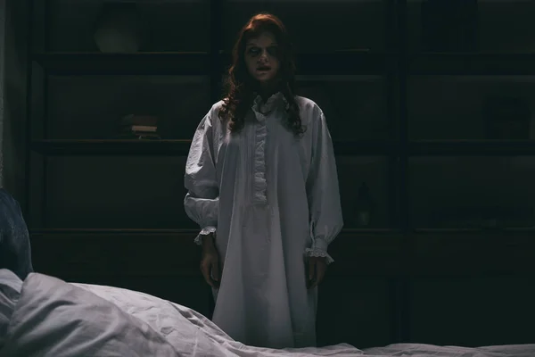 Mulher obcecada em camisola de pé perto da cama no quarto escuro — Fotografia de Stock