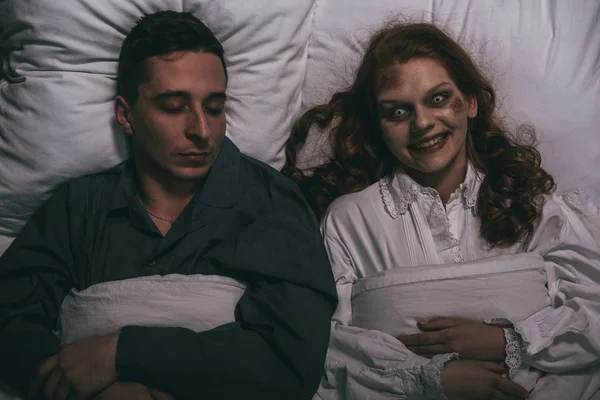 Вид сверху на криво улыбающуюся женщину-демона, лежащую в постели со спящим мужчиной — стоковое фото