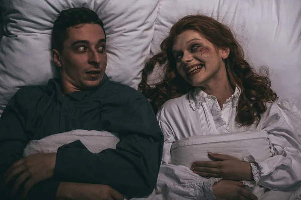 Draufsicht auf lächelnde Dämonin, die mit verängstigtem Mann im Bett liegt — Stockfoto