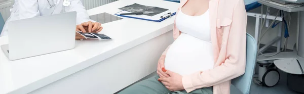 Panoramic shot of professional doctor showing ultrasound scan to young pregnant woman in clinic — Stock Photo