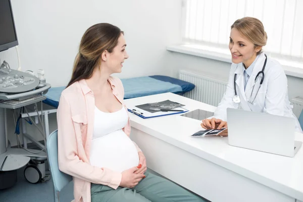 Sonriente doctor mostrando ultrasonido a joven embarazada en clínica - foto de stock