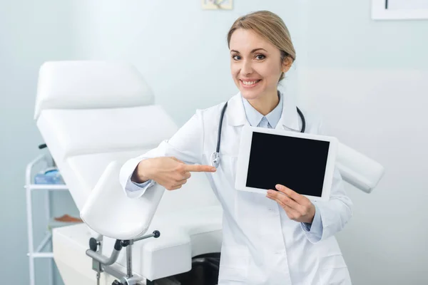 Ginecólogo sonriente apuntando a la tableta digital con pantalla en blanco en la clínica - foto de stock