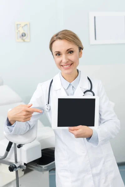 Улыбающаяся женщина-гинеколог указывает на цифровой планшет с пустым экраном в клинике — стоковое фото