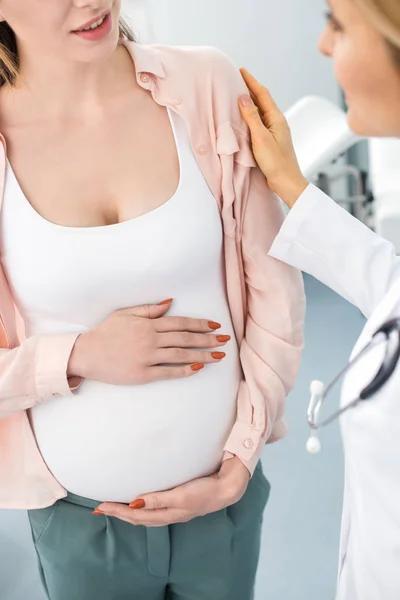 Cropped view of pregnant woman touching belly and having consultation with doctor in gynecological clinic — Stock Photo