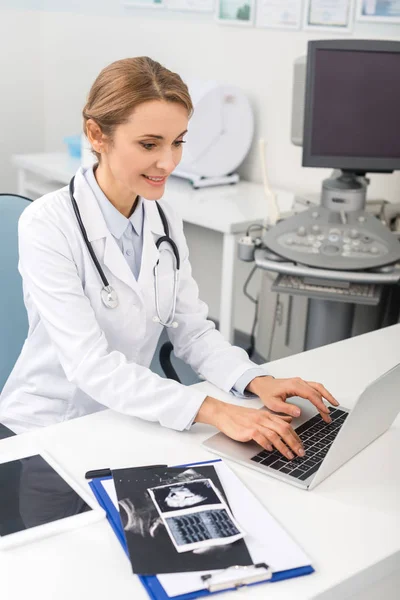 Professional female doctor working on laptop with ultrasound scans on table — Stock Photo