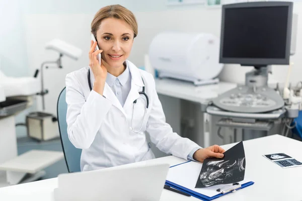 Médico sonriente profesional sosteniendo ultrasonido escaneo y hablando en el teléfono inteligente en la clínica - foto de stock