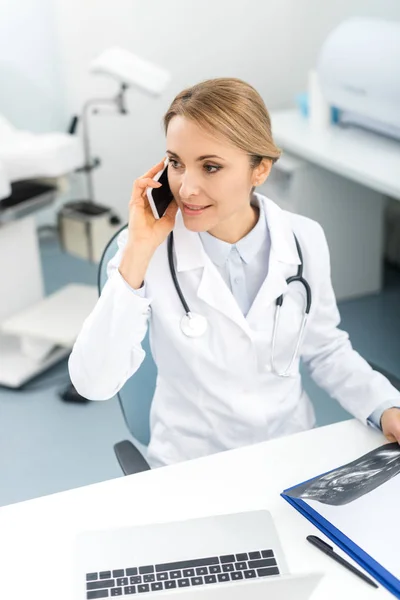 Hermoso médico sosteniendo ultrasonido escaneo y hablando en el teléfono inteligente en la clínica - foto de stock