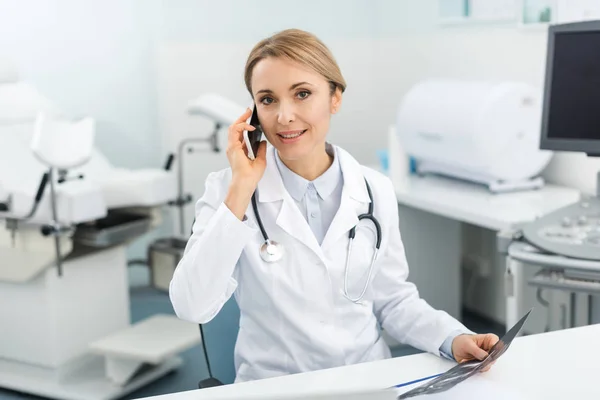 Doctora profesional sosteniendo ultrasonido escaneo y hablando en teléfono inteligente en la clínica - foto de stock