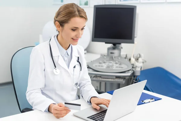 Médico sonriente usando computadora portátil y tarjeta de crédito en la clínica con escáner de ultrasonido - foto de stock