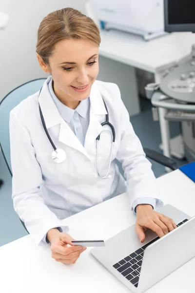 Hermoso médico usando el ordenador portátil y la tarjeta de crédito en la clínica - foto de stock