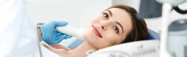 Panoramic shot of doctor examining thyroid of smiling patient with ultrasound scan in clinic — Stock Photo