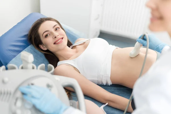 Врач осматривает живот улыбающейся беременной женщины с помощью УЗИ в клинике — стоковое фото