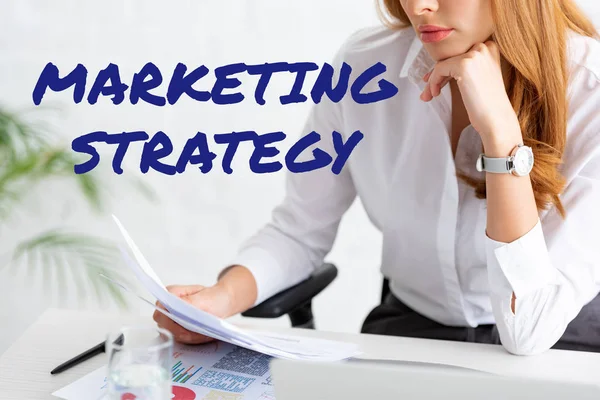 Обрезанный взгляд бизнесвумен, держащих досье рядом с графиками и ноутбуком на столе, иллюстрация маркетинговой стратегии — стоковое фото