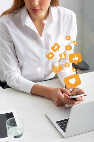 Vista recortada de una mujer de negocios sosteniendo un teléfono inteligente cerca de gadgets y un vaso de agua en la mesa, ilustración de redes sociales - foto de stock