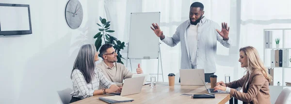 Panoramic shot of african american man talking with smiling colleagues during meeting in creative agency — Stock Photo