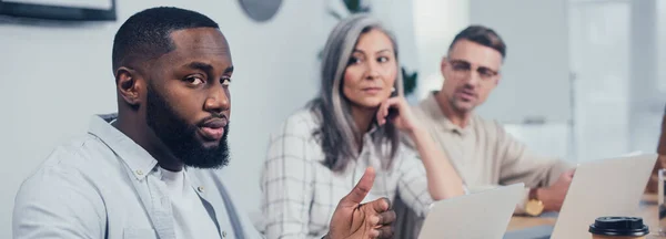 Panoramaaufnahme eines afrikanisch-amerikanischen mannes, der in die kamera schaut und seine kollegen, die ihn betrachten — Stockfoto