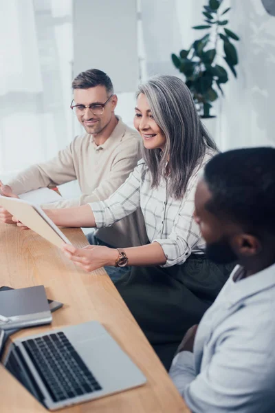 Smiling multicultural colleagues looking at folder during meeting in creative agency — Stock Photo