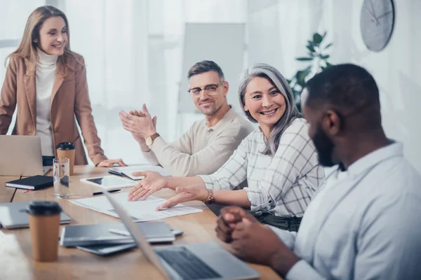 Smiling asian woman pointing with hands at folder and looking at african american colleague in creative agency — Stock Photo