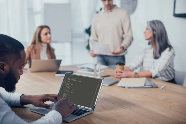 KYIV, UKRAINE - DECEMBER 6, 2019: selective focus of african american man using laptop with html code website and his colleagues talking on background — Stock Photo