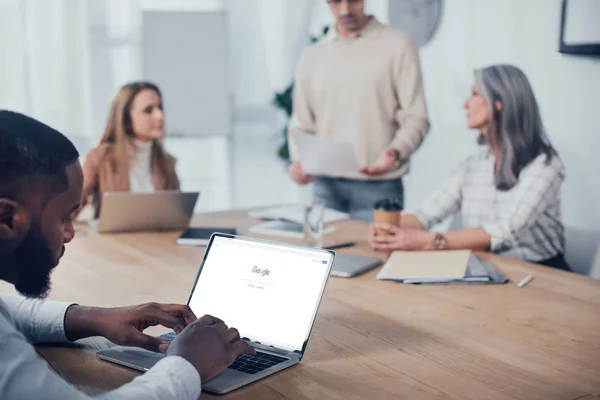 KYIV, UKRAINE - DECEMBER 6, 2019: selective focus of african american man using laptop with google and his colleagues talking on background — Stock Photo