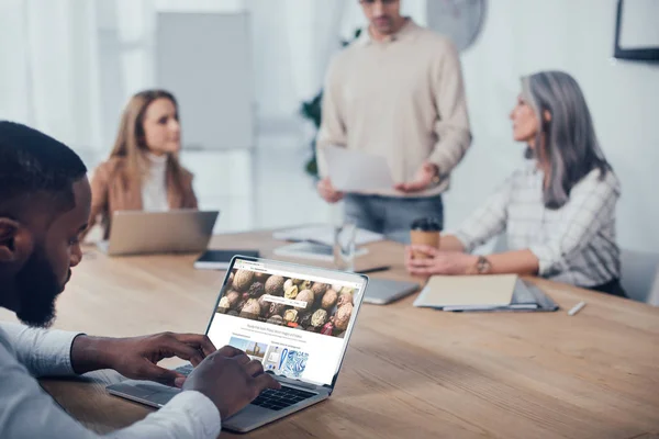 Kiew, Ukraine - 6. Dezember 2019: selektiver Fokus eines afrikanisch-amerikanischen Mannes, der einen Laptop mit Hintergrundfotos benutzt und seine Kollegen über Hintergründe sprechen — Stockfoto