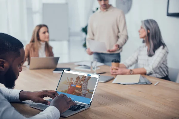 KYIV, UKRAINE - DECEMBER 6, 2019: selective focus of african american man using laptop with couchsurfing website and his colleagues talking on background — Stock Photo