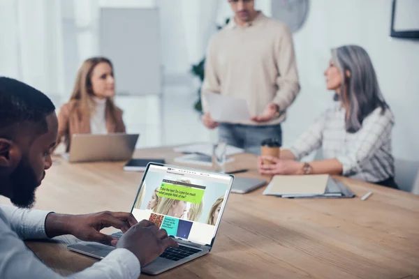 KYIV, UKRAINE - DECEMBER 6, 2019: selective focus of african american man using laptop with bbc website and his colleagues talking on background — Stock Photo