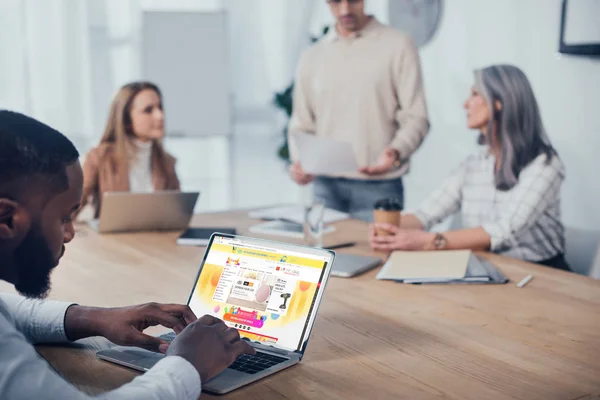 KYIV, UKRAINE - DECEMBER 6, 2019: selective focus of african american man using laptop with aliexpress website and his colleagues talking on background — Stock Photo