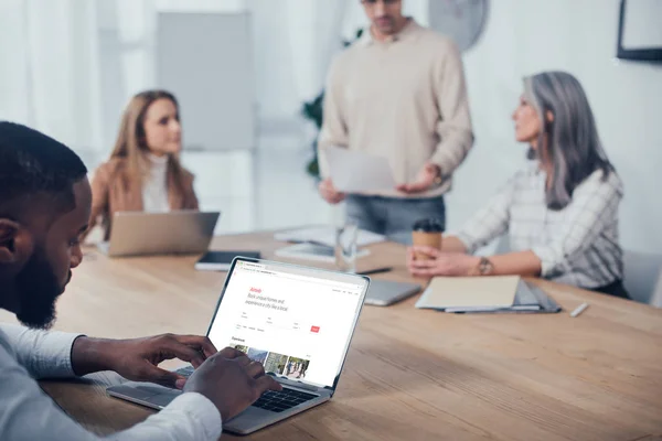 KYIV, UKRAINE - DECEMBER 6, 2019: selective focus of african american man using laptop with airbnb website and his colleagues talking on background — Stock Photo