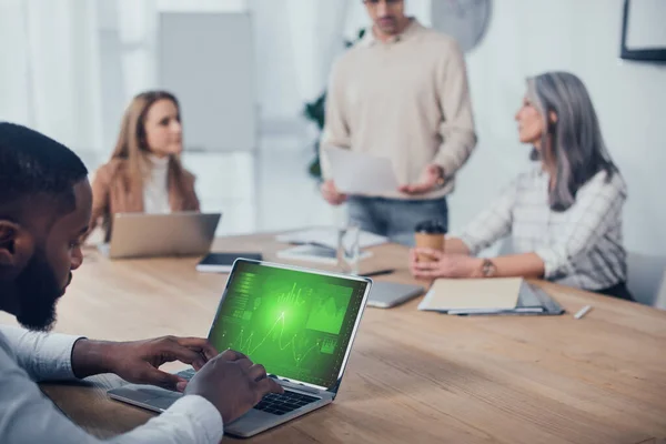 Foco seletivo do homem americano africano usando laptop com site de marketing de negócios e seus colegas falando em segundo plano — Fotografia de Stock