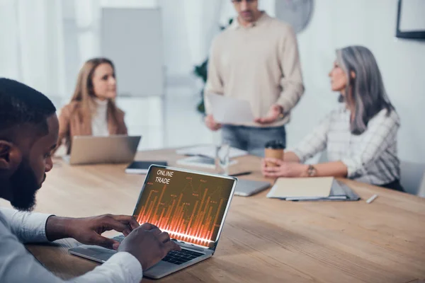 Enfoque selectivo del hombre afroamericano utilizando el ordenador portátil con el sitio web de comercio en línea y sus colegas hablando en segundo plano - foto de stock