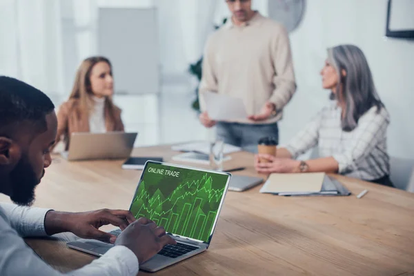 Enfoque selectivo del hombre afroamericano utilizando el ordenador portátil con el sitio web de comercio en línea y sus colegas hablando en segundo plano - foto de stock