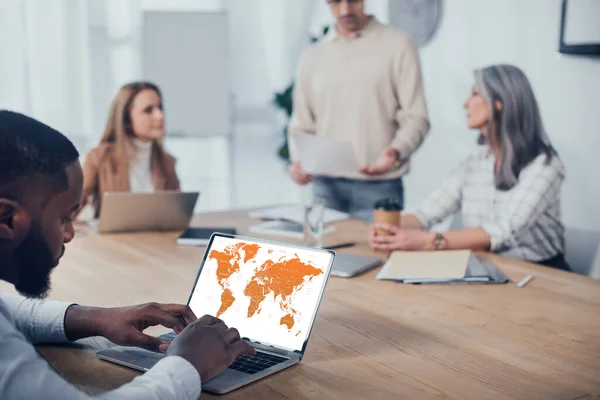 Enfoque selectivo del hombre afroamericano utilizando el ordenador portátil con mapa y sus colegas hablando sobre el fondo - foto de stock