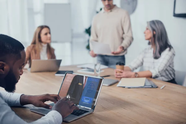 Messa a fuoco selettiva di uomo afro-americano utilizzando il computer portatile con il sito di prenotazione ei suoi colleghi che parlano in background — Foto stock