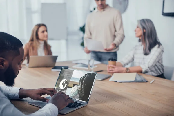 Selective focus of african american man using laptop with booking website and his colleagues talking on background — Stock Photo