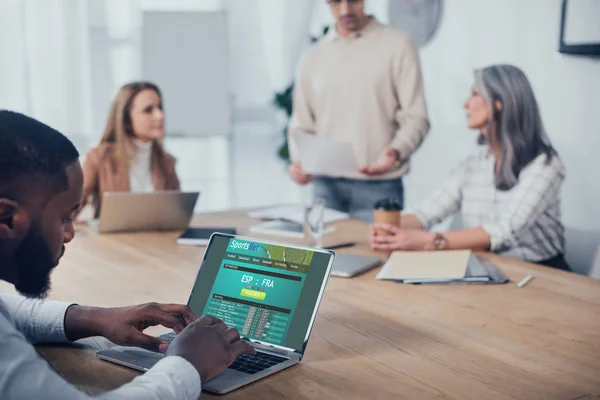 Selective focus of african american man using laptop with sports bet website and his colleagues talking on background — Stock Photo