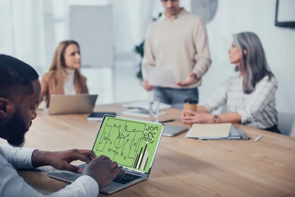 Selective focus of african american man using laptop with graphs and his colleagues talking on background — Stock Photo