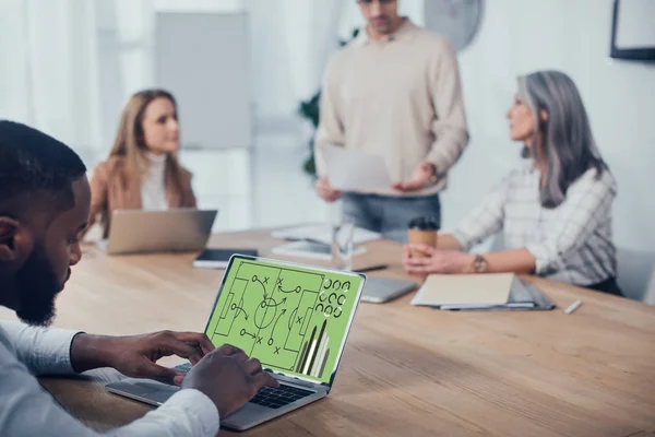 Messa a fuoco selettiva di uomo afro-americano utilizzando computer portatile con grafici ei suoi colleghi che parlano in background — Foto stock