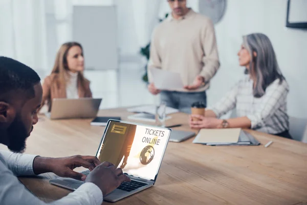 Foco seletivo do homem afro-americano usando laptop com bilhetes site on-line e seus colegas falando em segundo plano — Fotografia de Stock