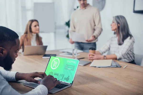 Selective focus of african american man using laptop with shopping website and his colleagues talking on background — Stock Photo