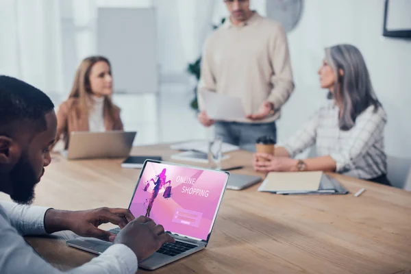Selective focus of african american man using laptop with online shopping website and his colleagues talking on background — Stock Photo