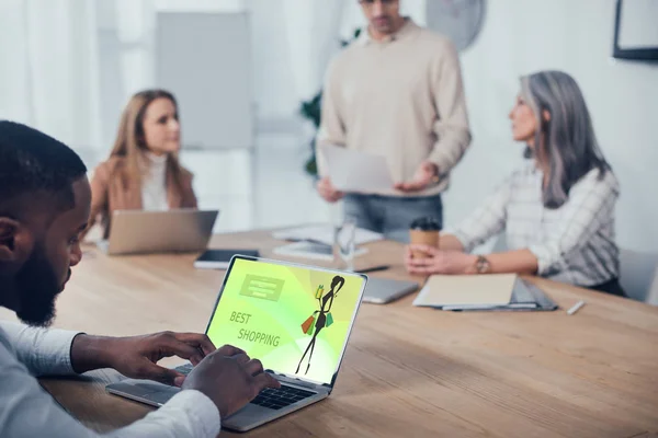 Selective focus of african american man using laptop with best shopping website and his colleagues talking on background — Stock Photo