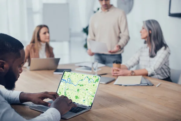 Selective focus of african american man using laptop with map website and his colleagues talking on background — Stock Photo