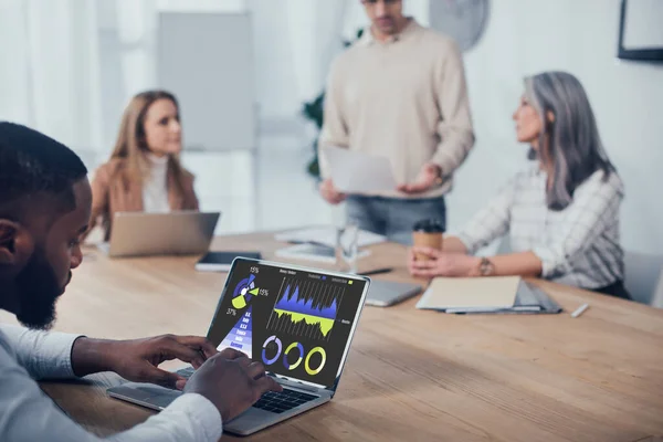 Selective focus of african american man using laptop with charts and graphs and his colleagues talking on background — Stock Photo