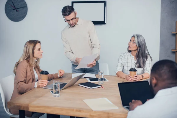 Messa a fuoco selettiva di colleghi multiculturali che fanno lavoro di ufficio e l'uomo afro-americano utilizzando il computer portatile su sfondo — Foto stock