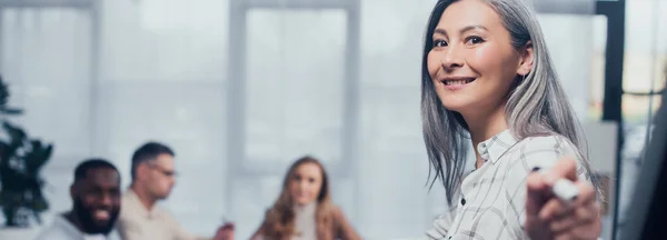 Plano panorámico de sonriente mujer de negocios asiática señalando con marcador y sus colegas multiculturales en el fondo - foto de stock