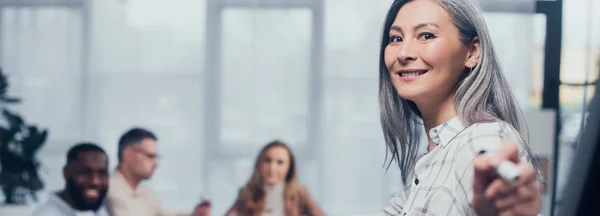 Panorâmica tiro de sorridente asiático empresária apontando com marcador e seus colegas multiculturais no fundo — Fotografia de Stock