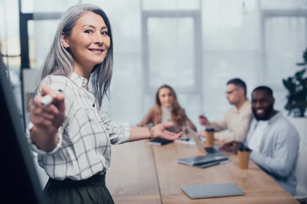 Selective focus of smiling asian businesswoman pointing with marker and her multicultural colleagues on background — Stock Photo